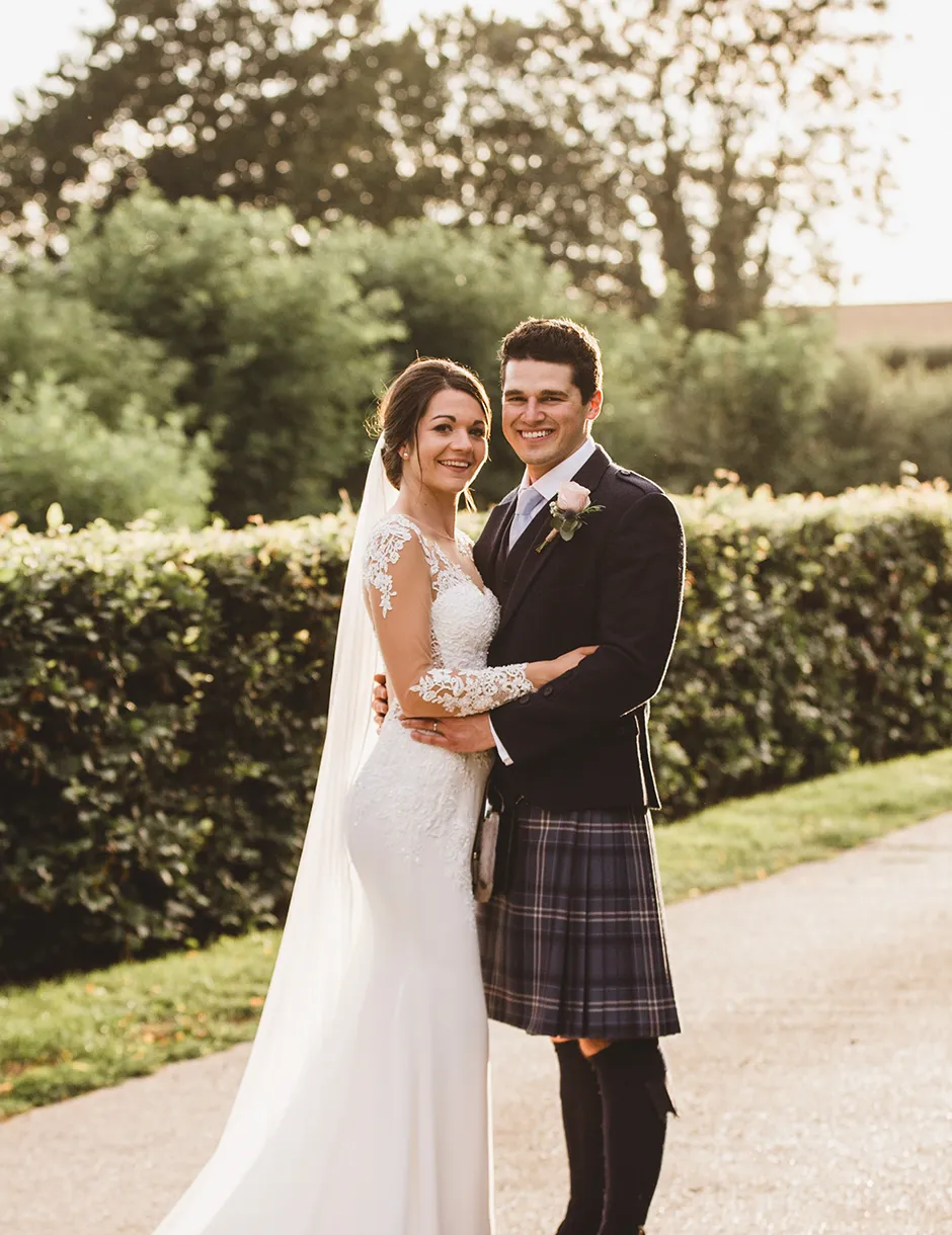 Young man and woman couple smiling and holding each other. Beautiful white wedding dress and navy blue kilt with a pink boutonniere.