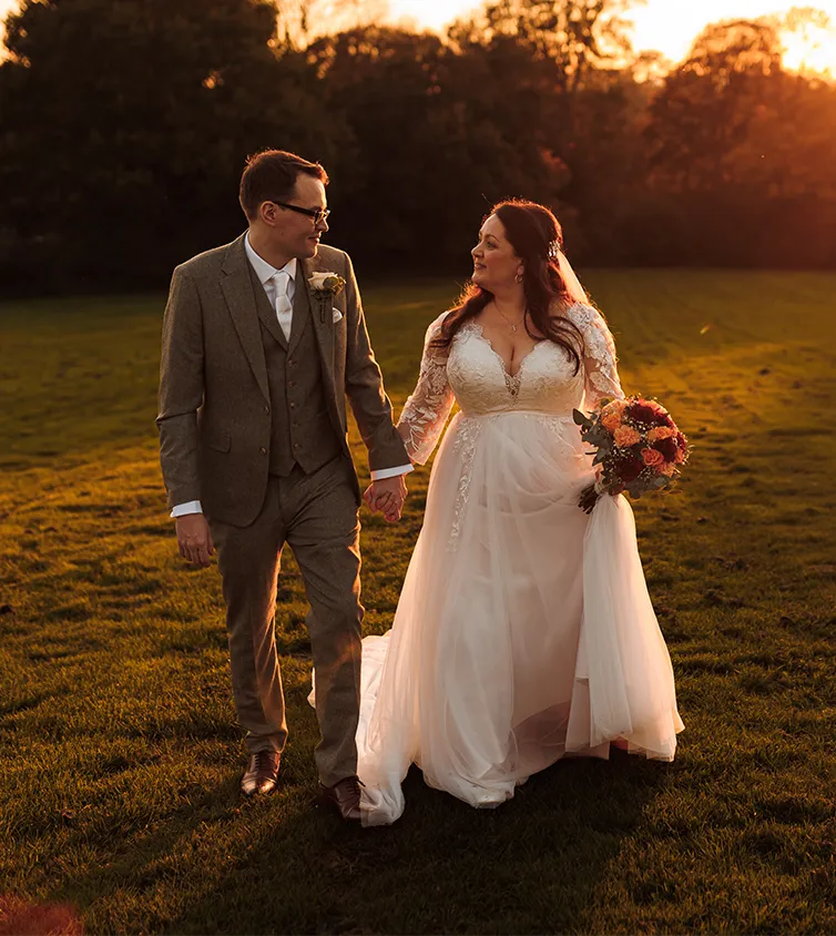 Young man and woman couple just married looking into each others eyes walking in a field with a sunset background holding flowers.