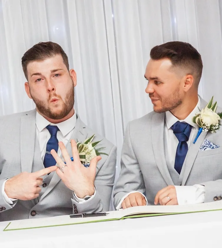 2 men just sitting at the registry table getting married, one man pointing at his wedding ring pouting wearing light grey suits