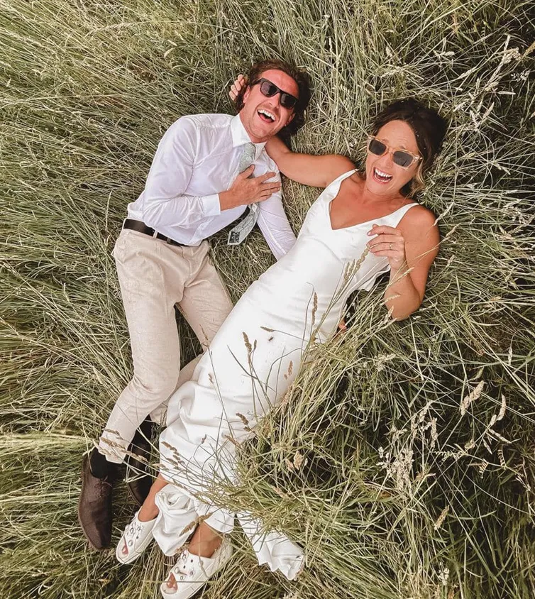 Birdseye view photo of a man and woman laying down in the grass. Wearing a smart shirt with beige trousers and a beautiful wedding dress.